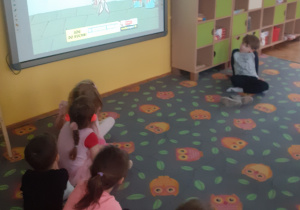 Dzieci z grupy fioletowej siedzą w półkolu na dywanie w sowy i oglądają filmik na temat dymów i ich źródeł na tablicy multimedialnej
